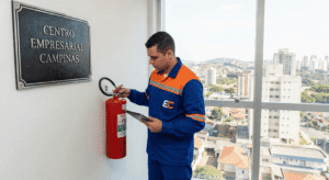 Técnico uniformizado de azul e laranja da empresa EC realizando a inspeção de extintores em um corredor do edifício Centro Empresarial Campinas, com uma ampla vista da cidade de Campinas ao fundo através da janela.